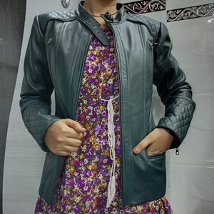May include: A dark green leather jacket featuring a mandarin collar with a snap button closure, quilted shoulder and sleeve panels, and zippered pockets. Underneath, a purple floral dress with a white drawstring waist detail is visible.