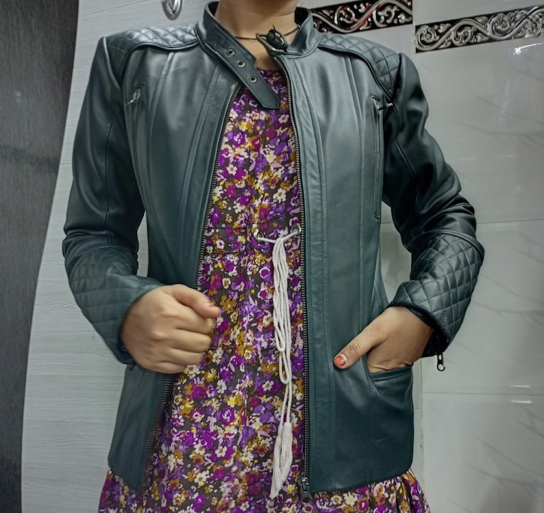 May include: A dark green leather jacket featuring a mandarin collar with a snap button closure, quilted shoulder and sleeve panels, and zippered pockets. Underneath, a purple floral dress with a white drawstring waist detail is visible.