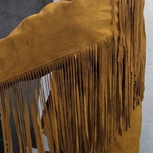 K&ouml;nnte beinhalten: Eine senfgelbe Jacke mit langen, d&uuml;nnen Fransen, die von der Schulter &uuml;ber den &Auml;rmel bis zum R&uuml;cken verlaufen. Das Kleidungsst&uuml;ck besteht aus einem strukturierten Material und weist sichtbare N&auml;hte entlang der Verbindungsstellen auf.