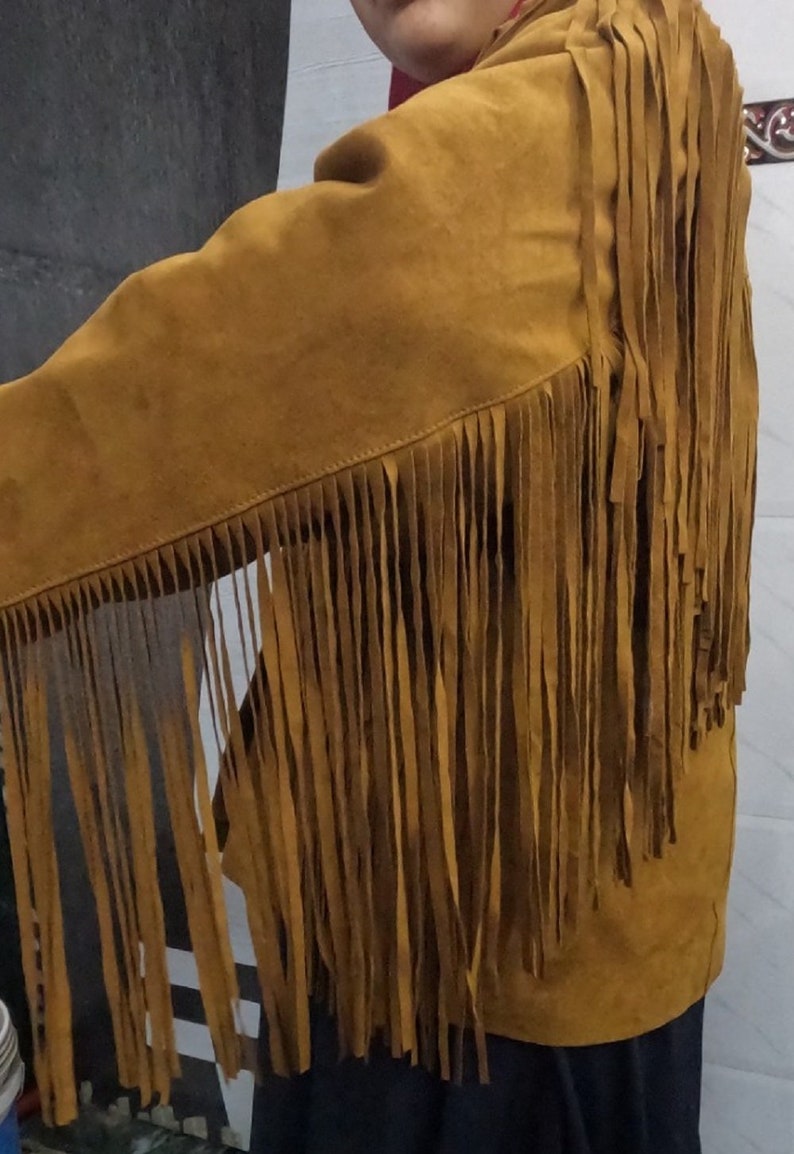 K&ouml;nnte beinhalten: Eine senfgelbe Jacke mit langen, d&uuml;nnen Fransen, die von der Schulter &uuml;ber den &Auml;rmel bis zum R&uuml;cken verlaufen. Das Kleidungsst&uuml;ck besteht aus einem strukturierten Material und weist sichtbare N&auml;hte entlang der Verbindungsstellen auf.