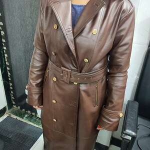 May include: A long, dark brown trench coat featuring a wide collar, double-breasted front with gold-toned buttons, and a matching belt with a buckle at the waist. The coat includes long sleeves with buttoned cuffs and vertical zippered side pockets.