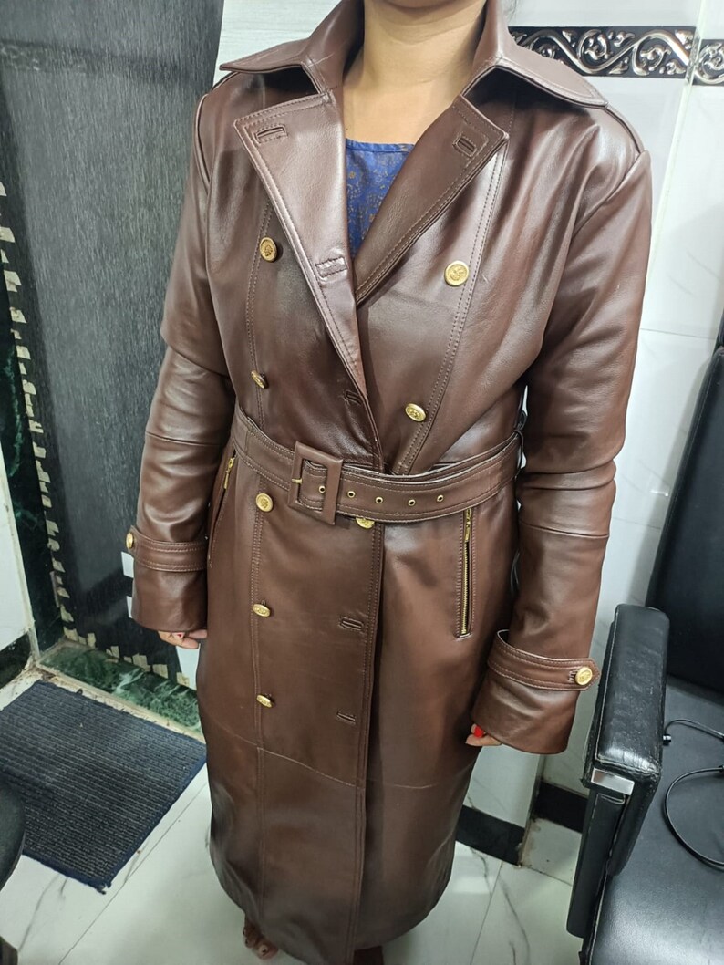 May include: A long, dark brown trench coat featuring a wide collar, double-breasted front with gold-toned buttons, and a matching belt with a buckle at the waist. The coat includes long sleeves with buttoned cuffs and vertical zippered side pockets.