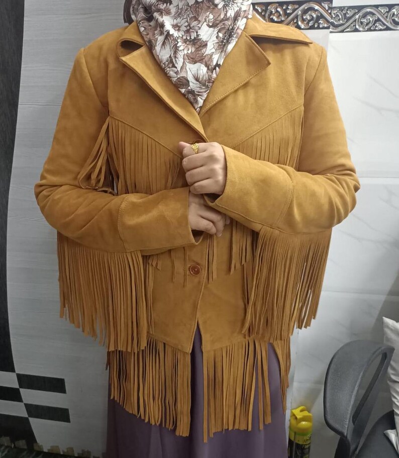 May include: A mustard yellow suede jacket featuring long fringe detailing along the chest, sleeves, and hemline. The jacket has a collared neckline, a button-front closure, and long sleeves. The material has a soft, matte texture throughout the garment.