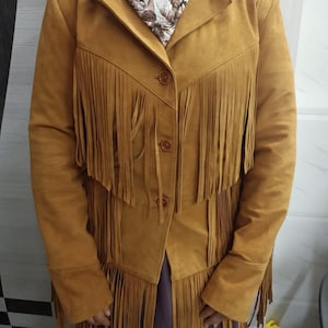 May include: A mustard yellow jacket featuring long fringe detailing across the chest and hemline. The jacket has a collared neckline, long sleeves, and three brown buttons down the front. A floral patterned scarf is worn around the neck.