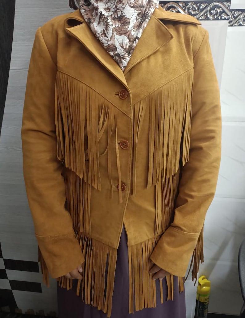 May include: A mustard yellow jacket featuring long fringe detailing across the chest and hemline. The jacket has a collared neckline, long sleeves, and three brown buttons down the front. A floral patterned scarf is worn around the neck.