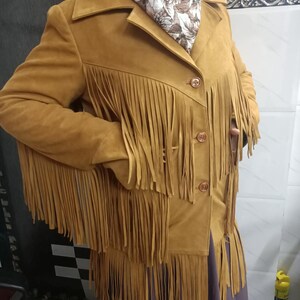 May include: A mustard yellow jacket featuring long fringe detailing across the chest, sleeves, and hem. The jacket has a collared neckline, three front buttons, and is worn over a dark purple garment with a floral patterned scarf at the neck.