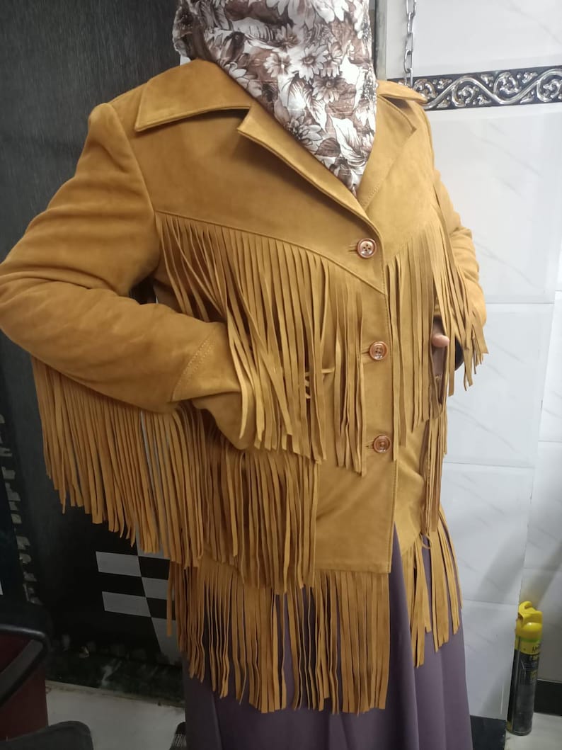 May include: A mustard yellow jacket featuring long fringe detailing across the chest, sleeves, and hem. The jacket has a collared neckline, three front buttons, and is worn over a dark purple garment with a floral patterned scarf at the neck.