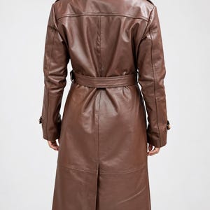 May include: A long, brown leather trench coat featuring a structured collar, shoulder epaulets, and long sleeves with buckled cuffs. The coat includes a matching waist belt with a buckle, a central back vent, and visible seam detailing throughout.