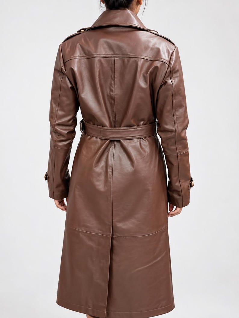 May include: A long, brown leather trench coat featuring a structured collar, shoulder epaulets, and long sleeves with buckled cuffs. The coat includes a matching waist belt with a buckle, a central back vent, and visible seam detailing throughout.