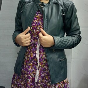 May include: A dark green leather jacket featuring quilted shoulder and sleeve panels, a stand-up collar with a buckle strap, and zippered pockets, worn over a purple floral print dress with a white drawstring waist detail.