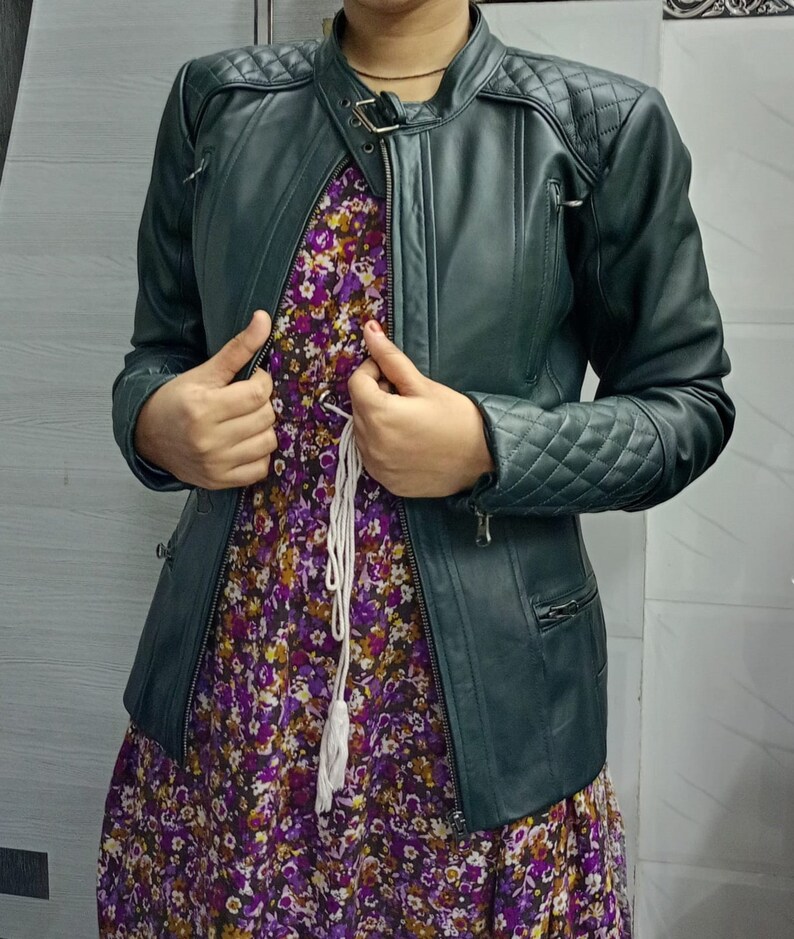 May include: A dark green leather jacket featuring quilted shoulder and sleeve panels, a stand-up collar with a buckle strap, and zippered pockets, worn over a purple floral print dress with a white drawstring waist detail.