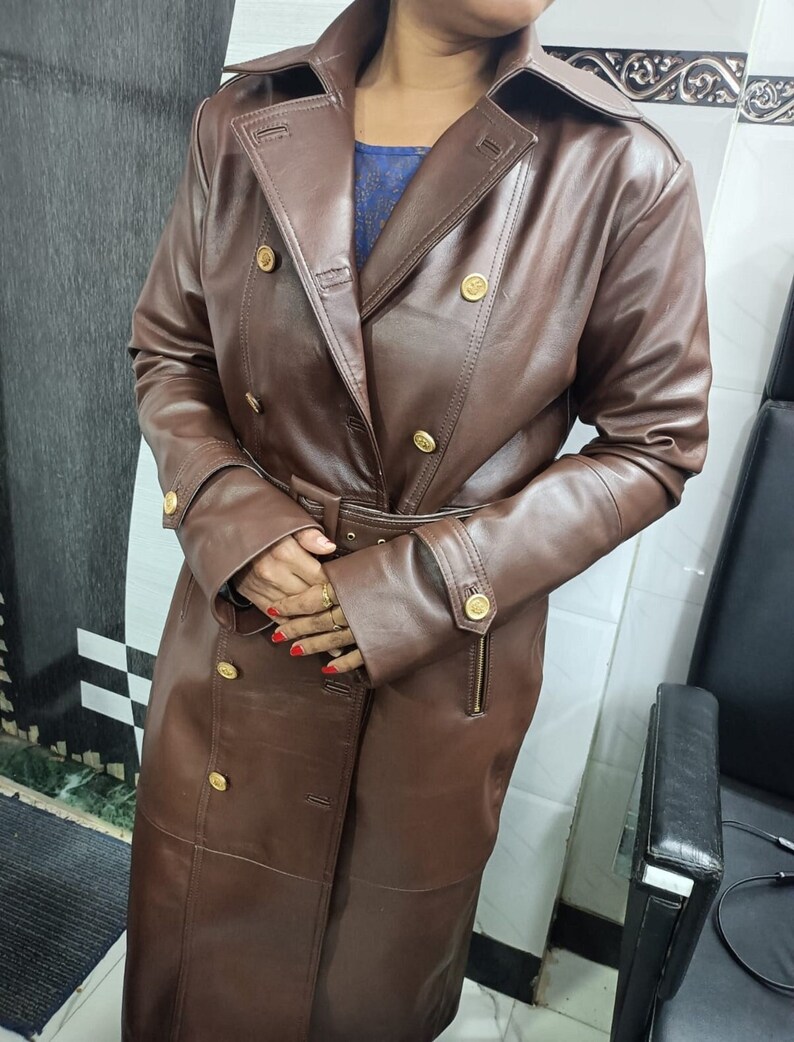 May include: A long, double-breasted brown coat featuring a wide collar, a matching belt with a buckle, and gold-toned buttons. The coat has long sleeves with buttoned cuffs and vertical zippered pockets on the front, providing a structured look.