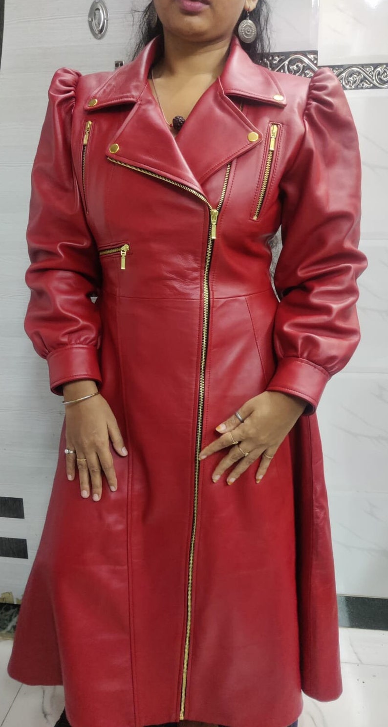 May include: A long, red leather coat featuring a wide collar with gold-tone snap buttons, a full-length front gold zipper, and three additional gold zippered pockets on the chest. The coat has long sleeves with gathered shoulders and cuffs.