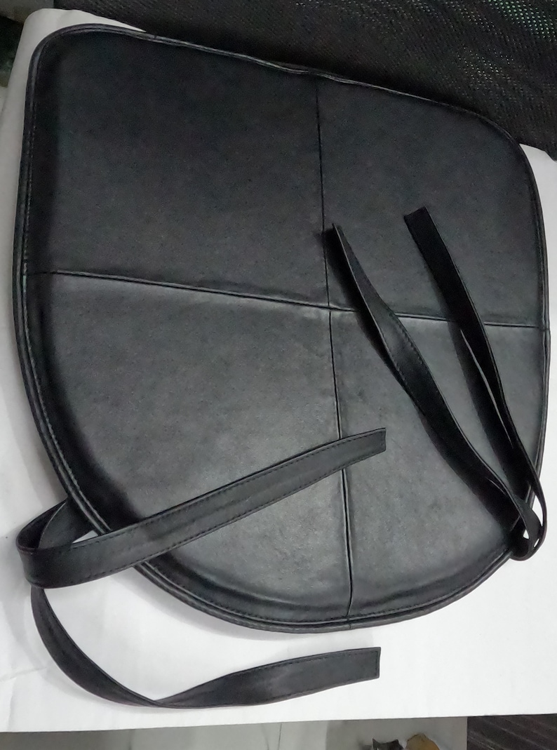May include: Black leather seat cushion with straps. The cushion is divided into four sections with stitched seams. The straps are long and thin, designed for securing the cushion.