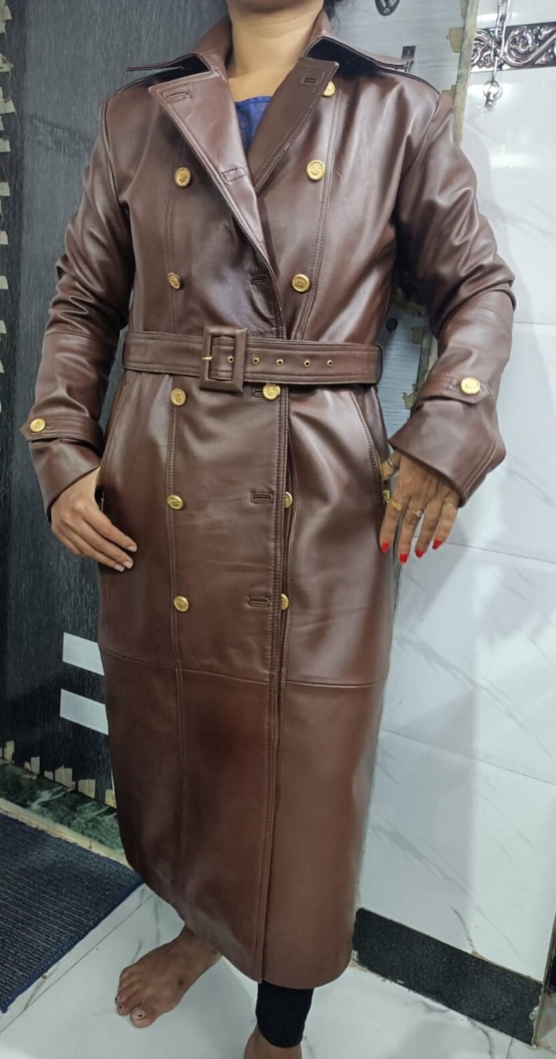 May include: A long, double-breasted brown coat featuring a wide collar, long sleeves with buttoned cuffs, and a matching belt with a rectangular buckle. The coat is adorned with multiple gold-toned buttons arranged in two vertical rows down the front.