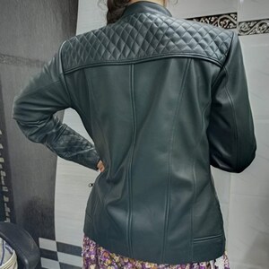 May include: A dark green jacket featuring a quilted diamond pattern across the upper back and shoulders, with matching quilted detailing on the sleeves. The jacket has a smooth finish, a stand-up collar, and vertical seam lines down the back.