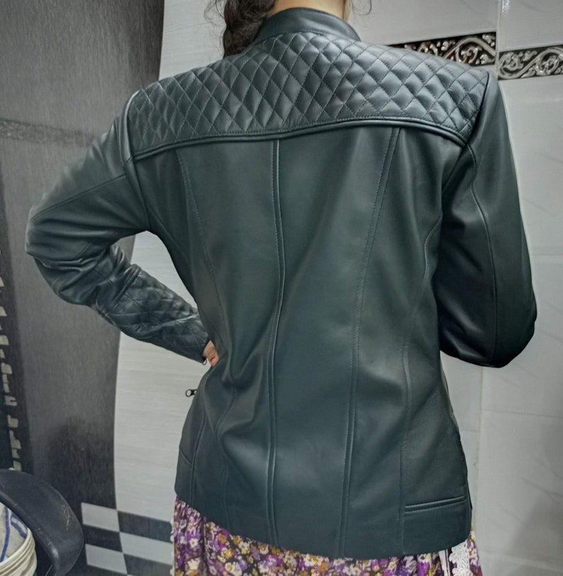 May include: A dark green jacket featuring a quilted diamond pattern across the upper back and shoulders, with matching quilted detailing on the sleeves. The jacket has a smooth finish, a stand-up collar, and vertical seam lines down the back.