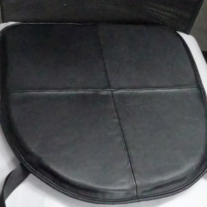 May include: Black leather seat cushion with a rounded front and stitched detailing. The cushion is divided into four sections by the stitching. Two black straps extend from the back of the cushion.