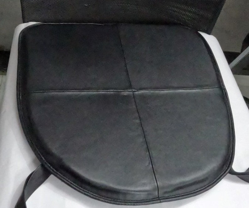 May include: Black leather seat cushion with a rounded front and stitched detailing. The cushion is divided into four sections by the stitching. Two black straps extend from the back of the cushion.