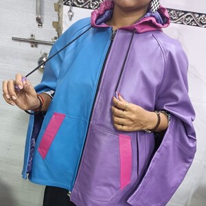 May include: A color-blocked zip-up jacket featuring a bright blue right side and a purple left side. The garment includes a purple hood, two front pockets with pink accents, and black drawstrings. The interior lining is a dark purple fabric.