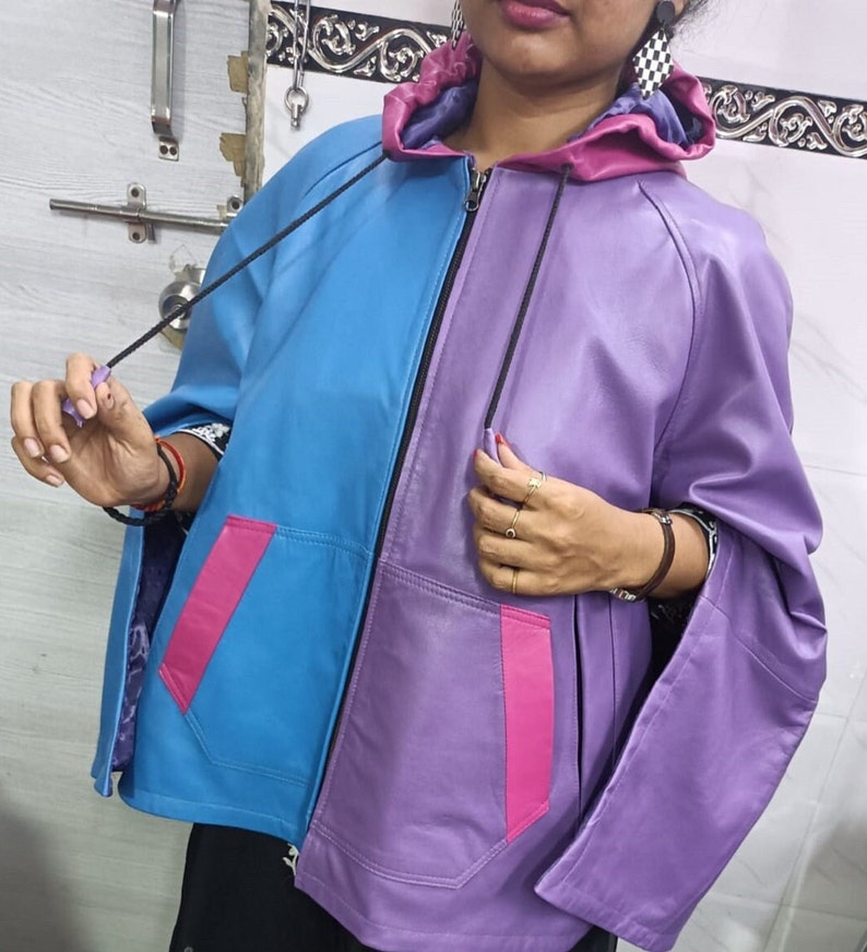 May include: A color-blocked zip-up jacket featuring a bright blue right side and a purple left side. The garment includes a purple hood, two front pockets with pink accents, and black drawstrings. The interior lining is a dark purple fabric.