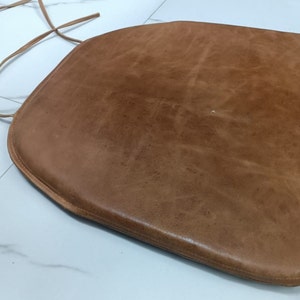 May include: A brown leather chair cushion with a tie closure. The cushion is made of soft, supple leather and has a slightly rounded shape.