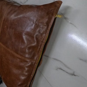 May include: A brown leather cushion with a yellow zipper. The cushion is made from soft, supple leather and has a smooth, even texture.