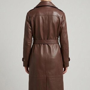 May include: A long, dark brown leather trench coat featuring a structured collar, shoulder epaulets, and a matching waist belt. The coat has long sleeves with buckled cuffs, a central back vent, and visible seam detailing throughout the garment.