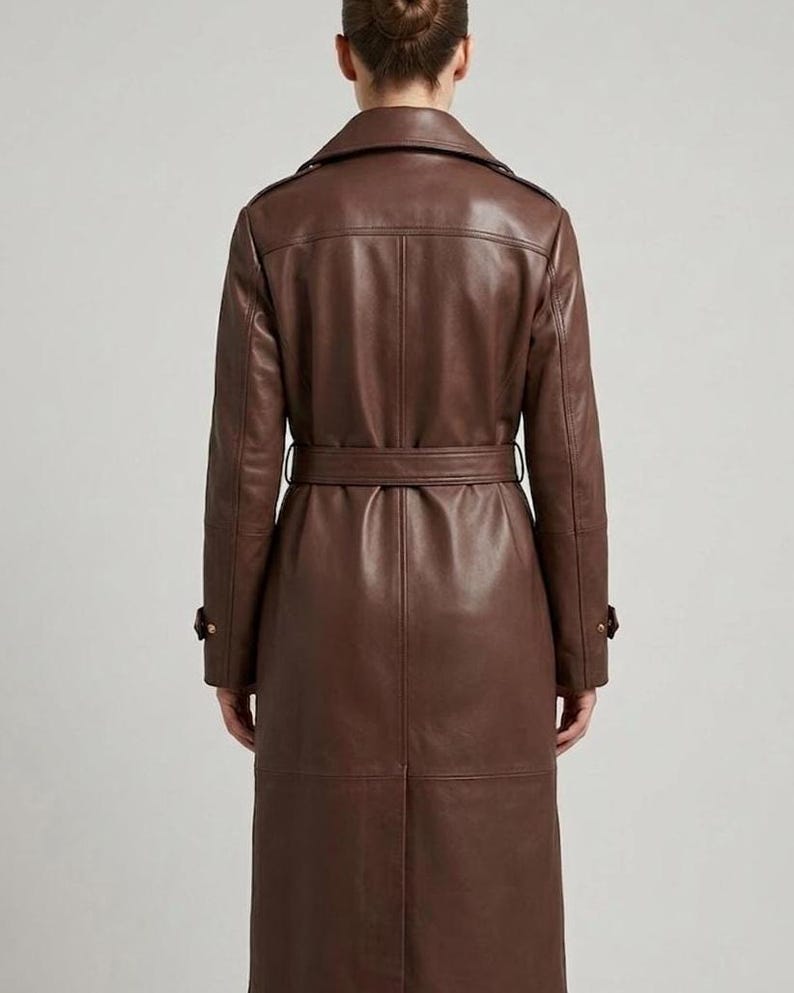May include: A long, dark brown leather trench coat featuring a structured collar, shoulder epaulets, and a matching waist belt. The coat has long sleeves with buckled cuffs, a central back vent, and visible seam detailing throughout the garment.