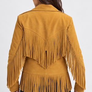 May include: A mustard yellow jacket featuring long fringe detailing across the back, shoulders, and hemline. The jacket has a collared neckline and long sleeves, with the fringe extending from the upper back yoke down to the lower hem.