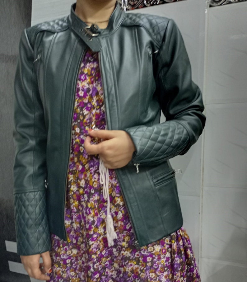 May include: A dark green jacket featuring a mandarin collar with a metal buckle closure, long sleeves with quilted diamond-patterned cuffs, and matching quilted detailing on the shoulders. The jacket is worn over a purple floral print dress.