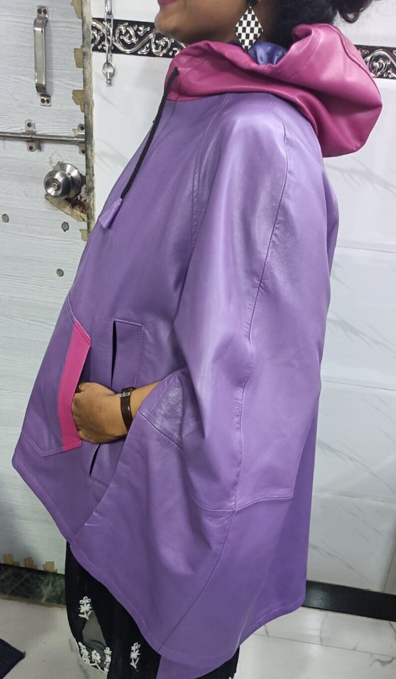May include: A lavender leather hooded jacket featuring a vibrant magenta hood lining and a matching magenta pocket trim. The jacket has wide sleeves, a smooth finish, and a black drawstring cord at the neck, providing a distinct color-blocked appearance.