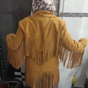 May include: A mustard yellow jacket featuring long fringe detailing across the back, shoulders, and sleeves. The garment has a wide collar and a V-shaped yoke design on the back, with the fringe extending from the yoke and the bottom hemline.
