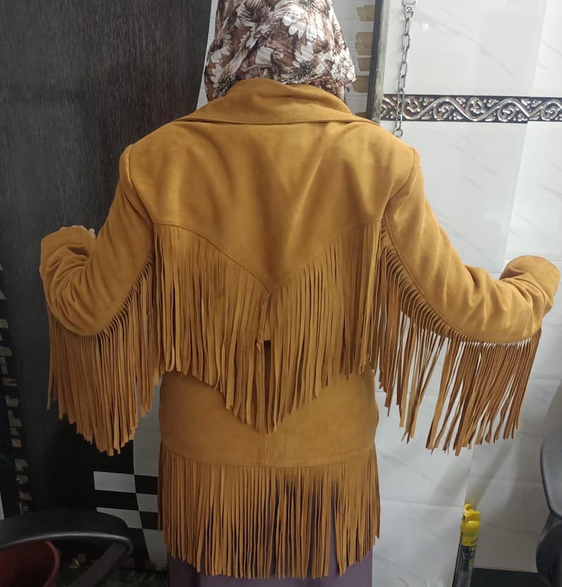 May include: A mustard yellow jacket featuring long fringe detailing across the back, shoulders, and sleeves. The garment has a wide collar and a V-shaped yoke design on the back, with the fringe extending from the yoke and the bottom hemline.
