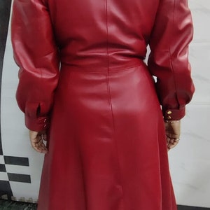 May include: A long-sleeved, knee-length red coat featuring a structured collar, puffed shoulders, and a cinched waist. The coat has a smooth finish, a central vertical seam down the back, and cuffs secured with small gold-toned buttons at the wrists.