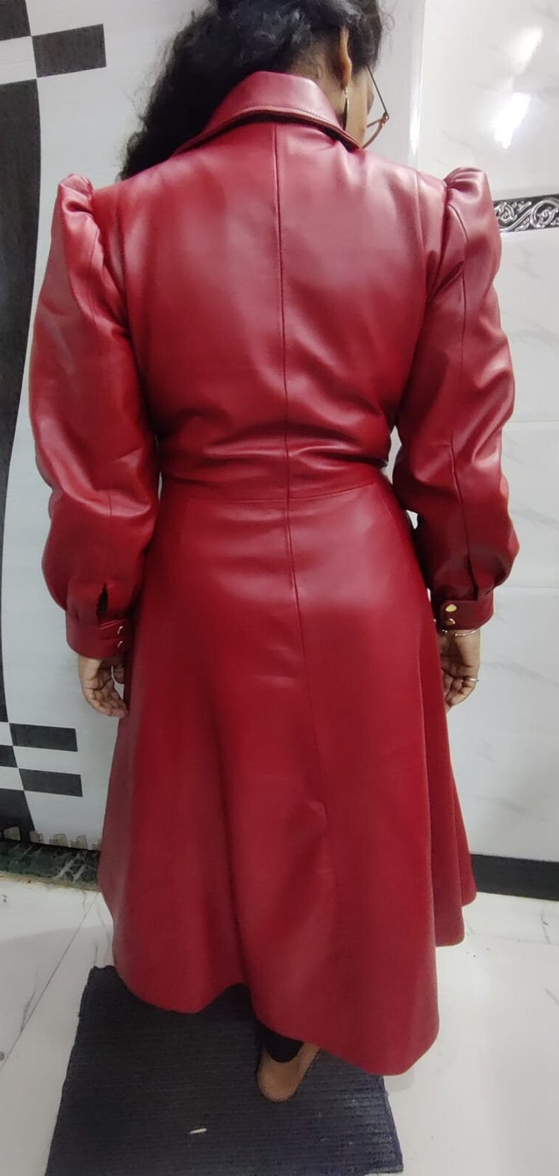 May include: A long-sleeved, knee-length red coat featuring a structured collar, puffed shoulders, and a cinched waist. The coat has a smooth finish, a central vertical seam down the back, and cuffs secured with small gold-toned buttons at the wrists.
