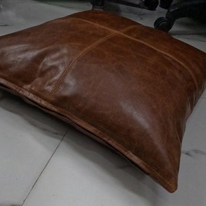 May include: A brown leather cushion with a square shape and a stitched design. The cushion is made of a soft, supple leather and has a slightly distressed look.