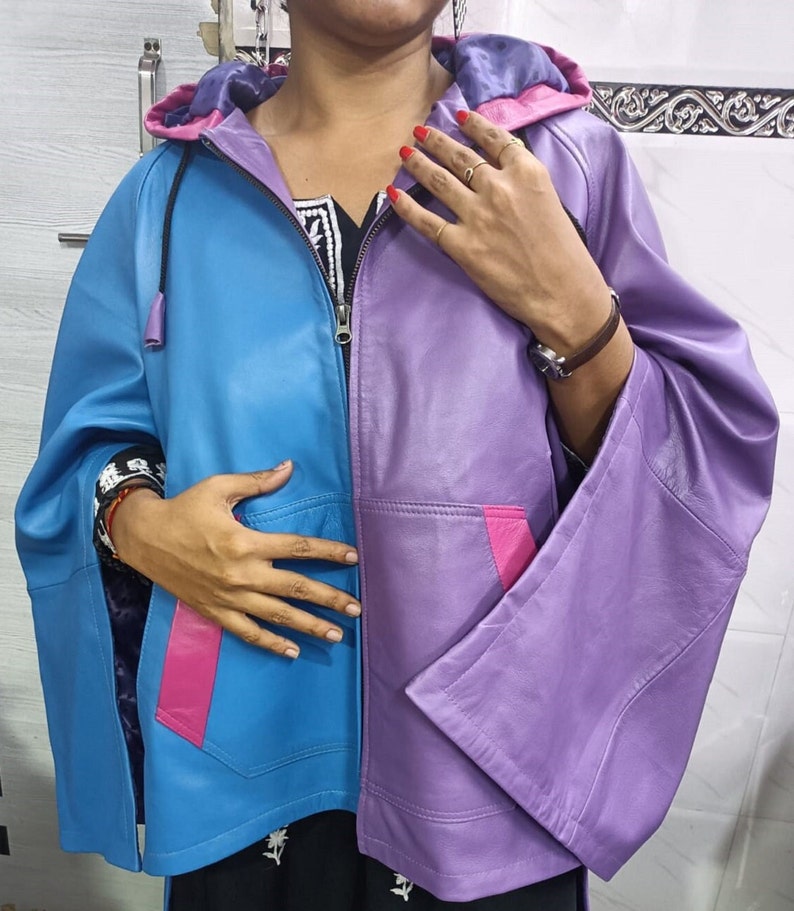 May include: A color-blocked hooded jacket featuring a bright blue right side and a purple left side. The jacket includes a front zipper, wide sleeves, and two front pockets accented with vibrant pink trim. The hood interior is lined in purple.
