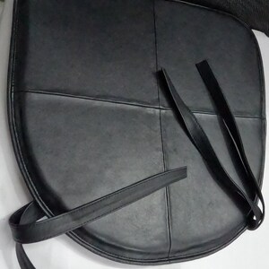 May include: Black leather seat cushion with straps. The cushion is rounded and divided into sections by stitched lines. The straps are long and thin, designed for securing the cushion. The cushion is likely for a chair.