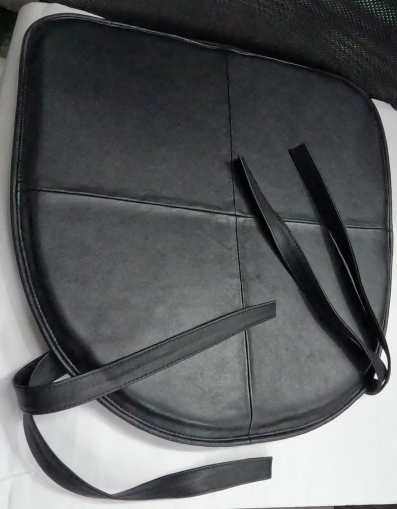 May include: Black leather seat cushion with straps. The cushion is rounded and divided into sections by stitched lines. The straps are long and thin, designed for securing the cushion. The cushion is likely for a chair.