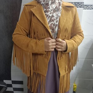 May include: A mustard yellow suede-textured jacket featuring long fringe detailing across the chest, sleeves, and hem. The jacket has a classic collar, a button-front closure, and long sleeves. It is worn over a floral patterned scarf and a solid purple garment.