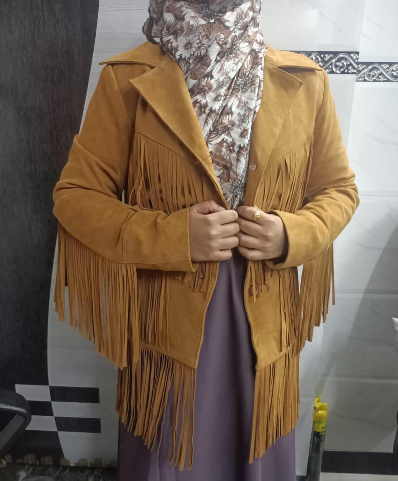 May include: A mustard yellow suede-textured jacket featuring long fringe detailing across the chest, sleeves, and hem. The jacket has a classic collar, a button-front closure, and long sleeves. It is worn over a floral patterned scarf and a solid purple garment.