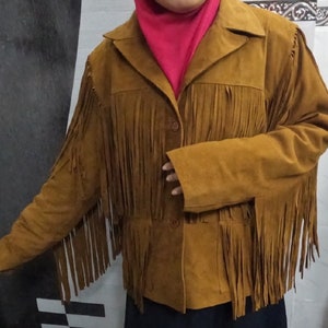 K&ouml;nnte beinhalten: Eine hellbraune Wildlederjacke mit langen Fransen an Brust, R&uuml;cken und &Auml;rmeln. Die Jacke verf&uuml;gt &uuml;ber einen klassischen Kragen, eine Knopfleiste auf der Vorderseite und lange &Auml;rmel. Das weiche, strukturierte Material sorgt f&uuml;r einen Look.