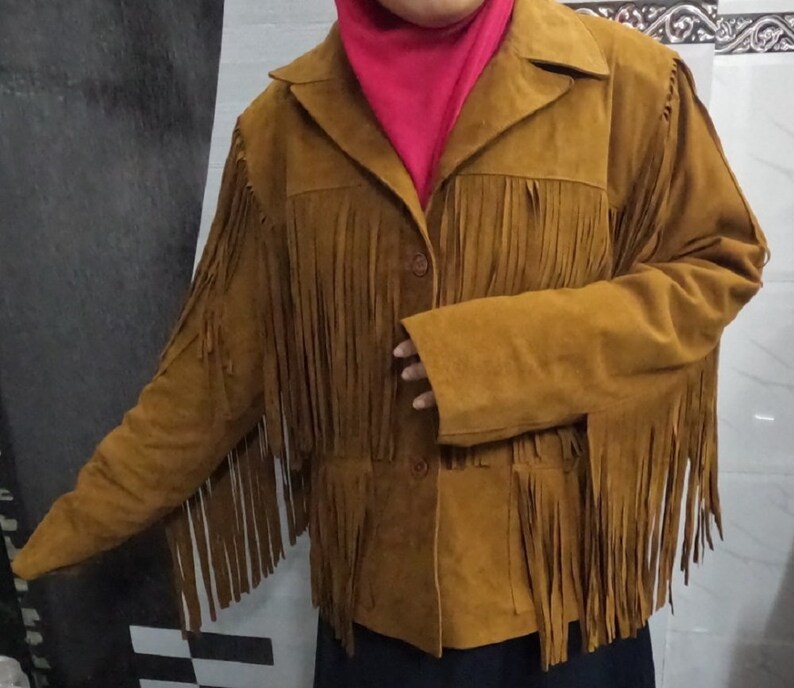 K&ouml;nnte beinhalten: Eine hellbraune Wildlederjacke mit langen Fransen an Brust, R&uuml;cken und &Auml;rmeln. Die Jacke verf&uuml;gt &uuml;ber einen klassischen Kragen, eine Knopfleiste auf der Vorderseite und lange &Auml;rmel. Das weiche, strukturierte Material sorgt f&uuml;r einen Look.