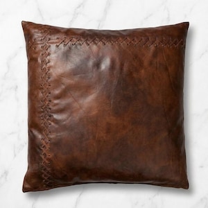 Puede incluir: Un cojín cuadrado hecho de cuero marrón oscuro. La superficie frontal presenta un patrón decorativo de punto de cruz a lo largo de los bordes superior e izquierdo, creado con finas tiras de cuero pasadas por pequeños agujeros perforados.