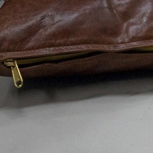 May include: A brown leather cushion with a gold zipper. The cushion is lying on a white surface.