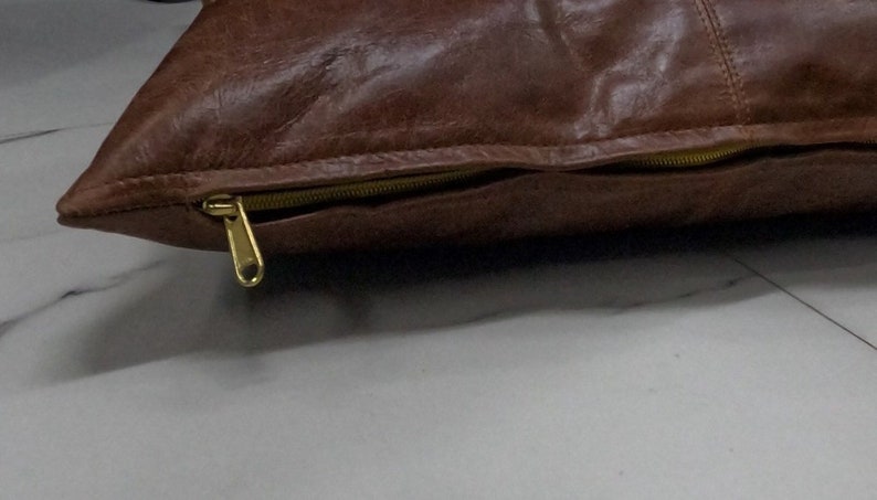 May include: A brown leather cushion with a gold zipper. The cushion is lying on a white surface.