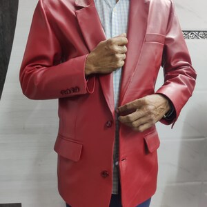 Może przedstawiać: Żywy czerwony sk&oacute;rzany blazer z klasycznym zapięciem na dwa guziki i klapami z wycięciami. Blazer posiada dwie przednie kieszenie i mankiety zapinane na guziki. Kurtka jest noszona na jasnoniebieskiej i białej koszuli w kratę oraz niebieskich spodniach.
