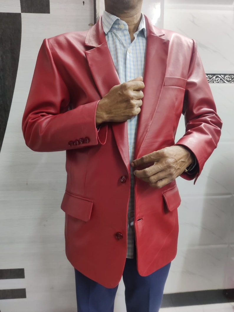 Może przedstawiać: Żywy czerwony sk&oacute;rzany blazer z klasycznym zapięciem na dwa guziki i klapami z wycięciami. Blazer posiada dwie przednie kieszenie i mankiety zapinane na guziki. Kurtka jest noszona na jasnoniebieskiej i białej koszuli w kratę oraz niebieskich spodniach.