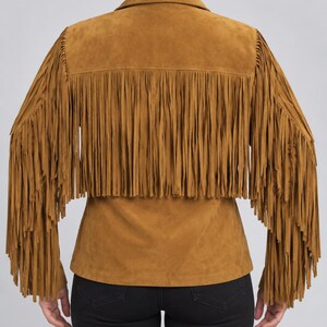 K&ouml;nnte beinhalten: Eine hellbraune Wildlederjacke mit langen Fransen am R&uuml;cken und an den &Auml;rmeln. Die Jacke hat einen klassischen Kragen und einen geraden Saum, was ihr durch die weiche, matte Oberfl&auml;che und die einheitliche erdige Farbe eine Textur verleiht.